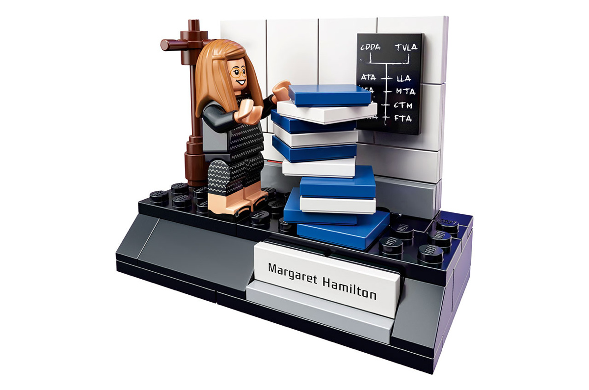 Figurine Lego de Margaret Hamilton devant une pile de livres bleus et blancs, avec un tableau noir et sa plaque nominative