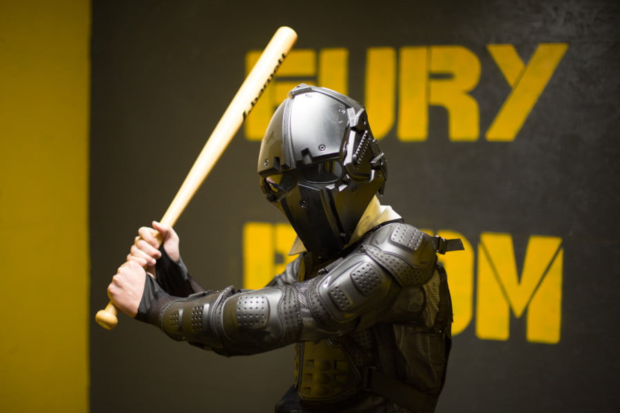 Combattant masqué en armure, casque intégral, brandissant une batte dans une fury room