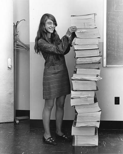 Margaret Hamilton souriante, posant à côté d'une pile de listings du code d'Apollo aussi haute qu'elle